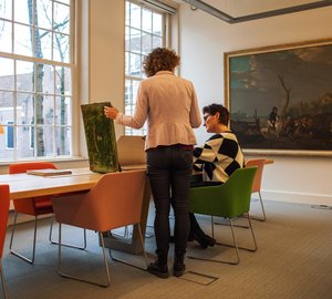 In de Studiezaal Regionaal Archief Dordrecht zit een vrouw aan tafel. De staande vrouw naast haar toont haar een groot boek.